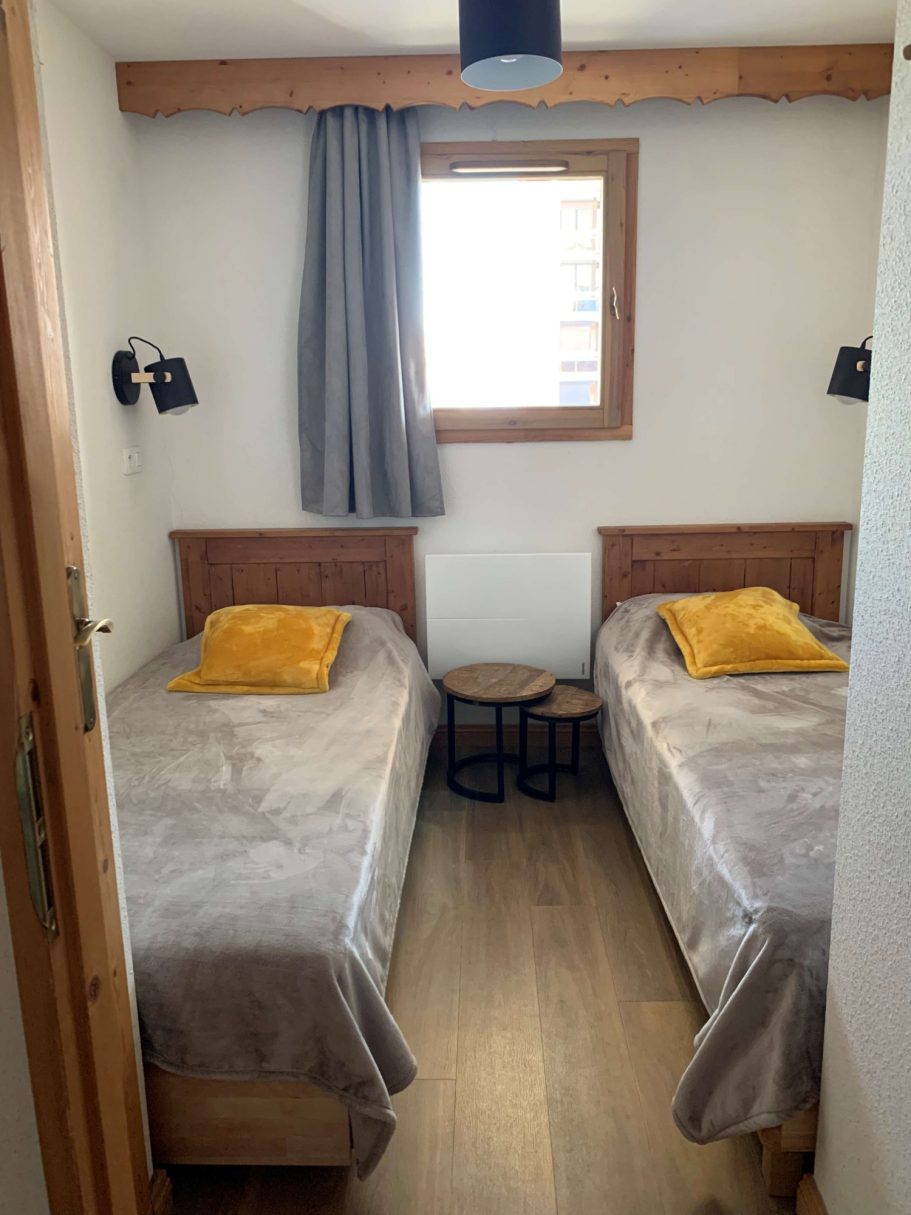 Chambre avec deux lits simples, rideaux gris et coussins jaunes, espace lumineux.